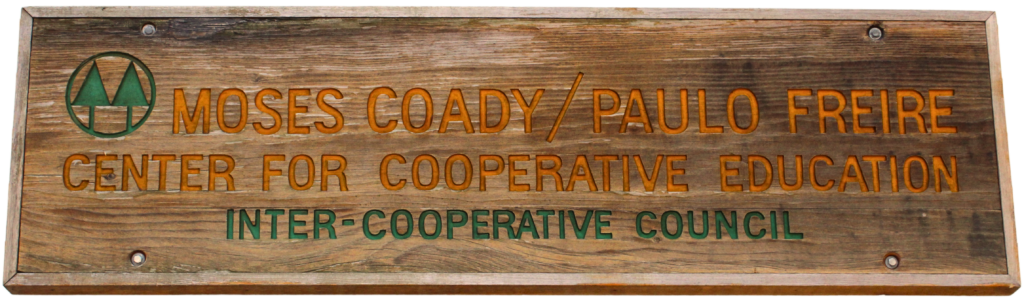 Sign for the Moses Coady/Paulo Freire Center for Cooperative Education at the Inter-Cooperative Council. Painted with green and orange lettering on a worn plank of wood and screwed to the front of the aforementioned building.
